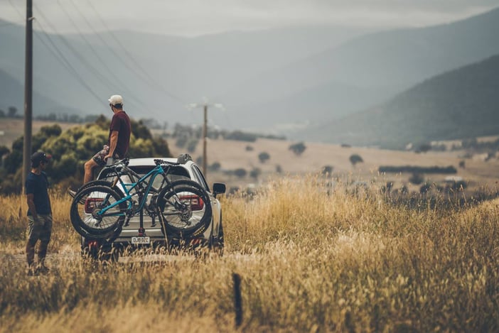 Two people are standing with their parked car taking in a summer valley view. There are two mountain bikes on the rear bike rack and golden grass in the foreground