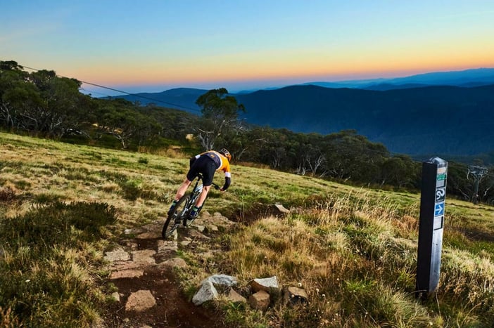 A rider is standing on the pedals of their mountain bike heading downhill with a sweeping alpine view ahead of them. There is a trail marker showing the way to Copperhead trail. The rider is in lycra ride gear and a helmet