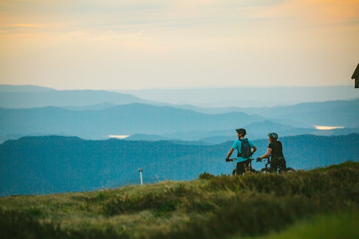 Two riders are stopped on their mountain bikes taking in a wide view across the high country ranges. There are lush green plants and the layers of mountains in the background are different shades of blue with patches of mist